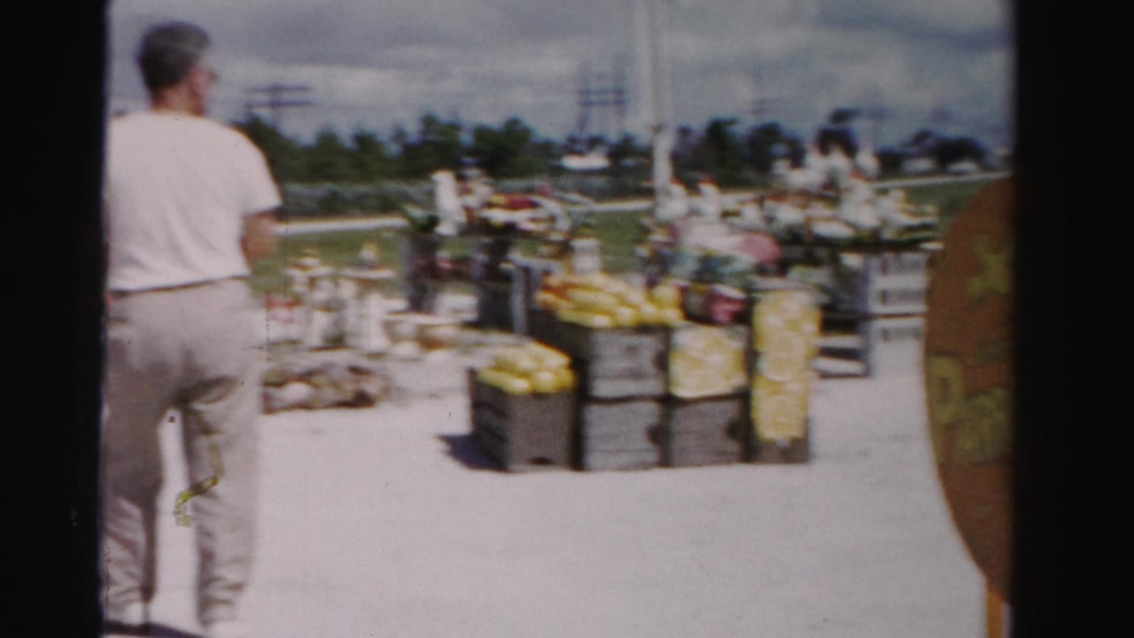 Man in a white T-shirt approaches a roadside produce stand with crates and signs ahead.