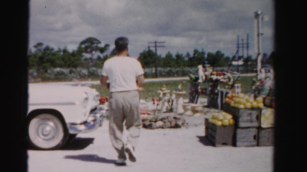 Man walks past a white vintage car toward a roadside stand with produce crates.