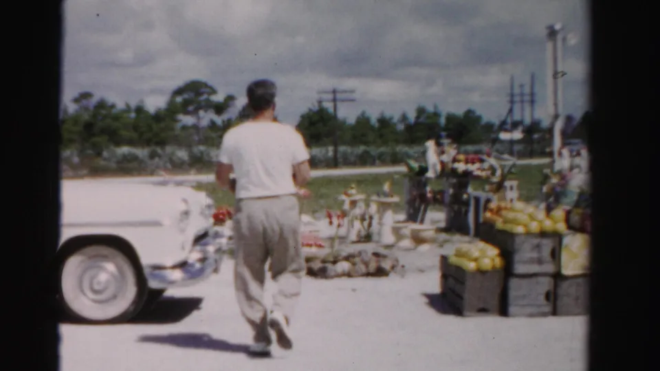 In 1955 Dade County, Florida, a man passes a white vintage car while approaching a roadside produce stand with stacked crates.