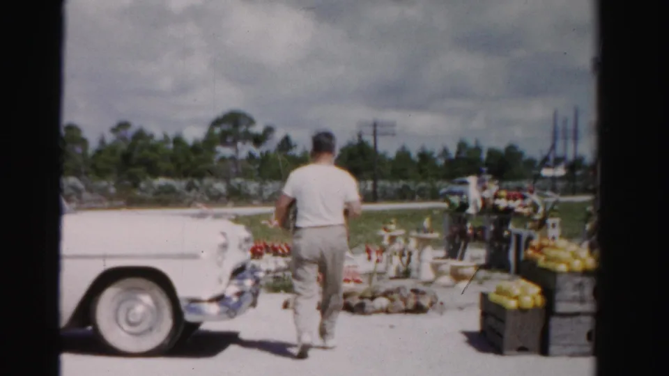 A 1955 Dade County home-movie frame shows the man near the front of a white car, facing produce crates and roadside display items.