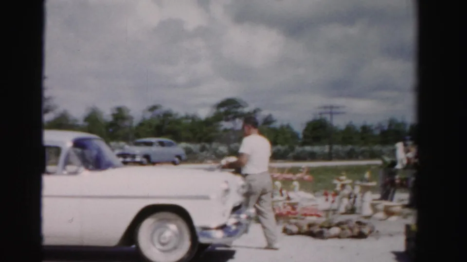 In 1955 Dade County, Florida footage, the white vintage car sits beside a roadside stand while the man stands near produce displays.