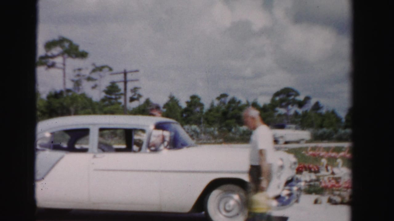1955 Dade County frame of a man carrying a yellow bag and oranges beside a two-tone sedan near a roadside stand.