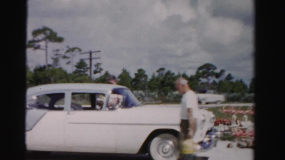 1955 Dade County frame of a man carrying a yellow bag and oranges beside a two-tone sedan near a roadside stand.