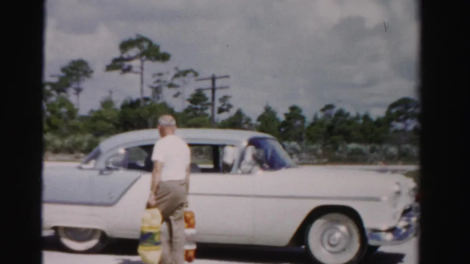 1955 Dade County home-movie frame showing a man holding groceries beside the passenger side of a classic sedan.
