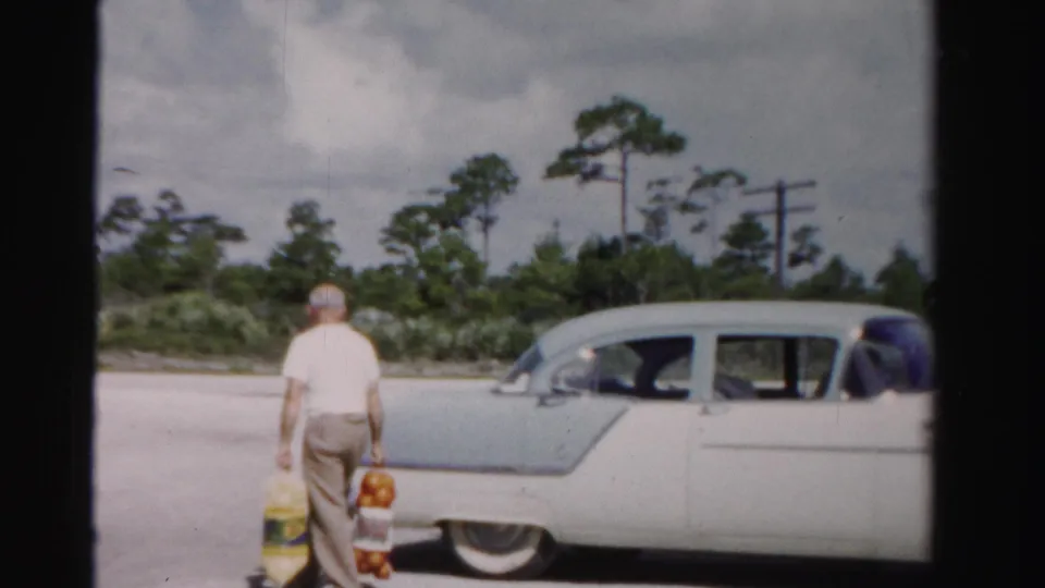 1955 Dade County frame of a man seen from behind carrying groceries toward a two-tone sedan under a cloudy sky.
