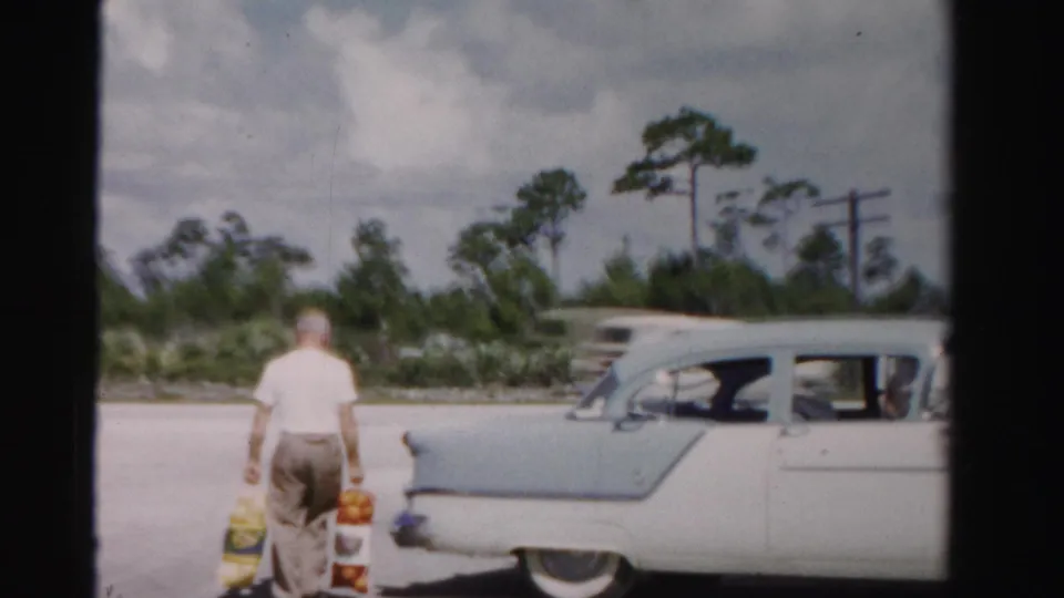 In 1955 Dade County, a man carries bags of fruit toward a pale blue-and-white sedan beside a road with trees and utility poles.