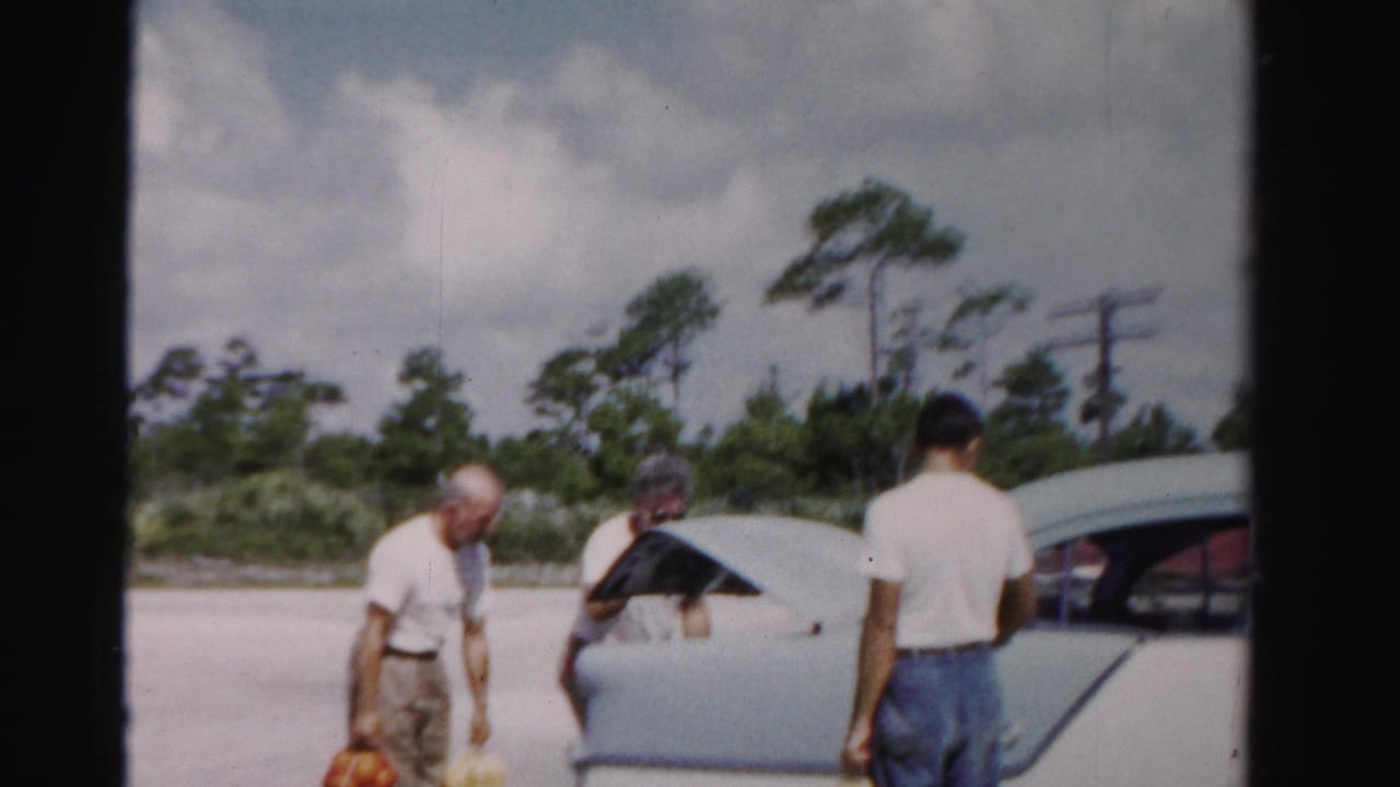 Three men stand by a pale sedan as the trunk is opened for groceries.