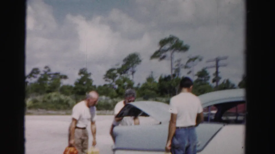 Three adult men gather at a pale mid-century sedan in Dade County, Florida, with grocery bags visible as the trunk is opened.