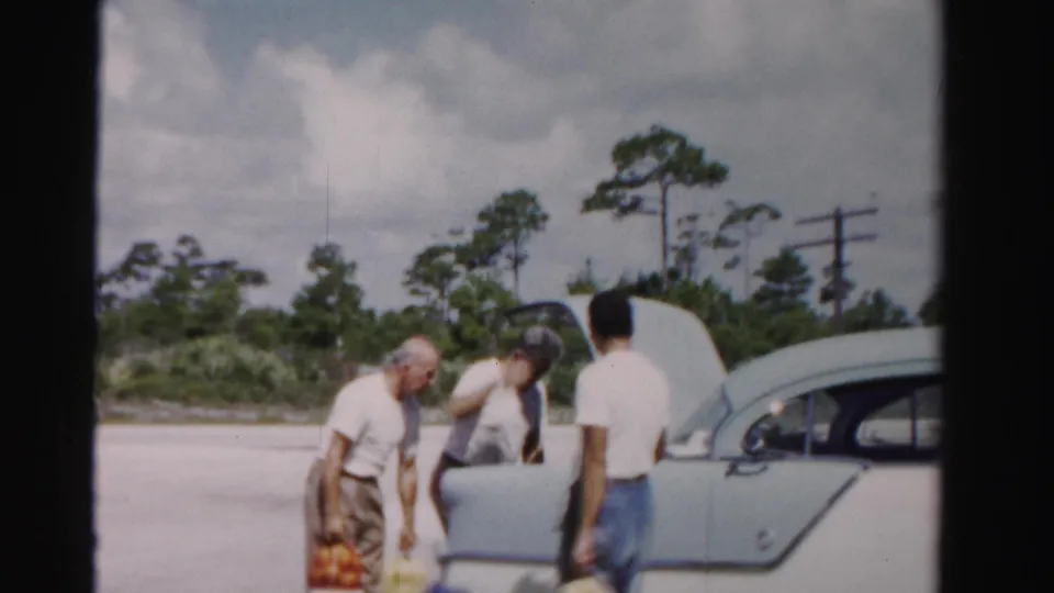 In the Dade County home-movie scene, two men lean toward the open trunk while another stands beside the pale two-tone sedan.
