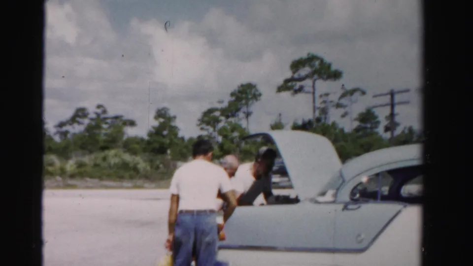 Three men load groceries into the open trunk of a pale 1950s sedan in a sunny Dade County roadside setting.
