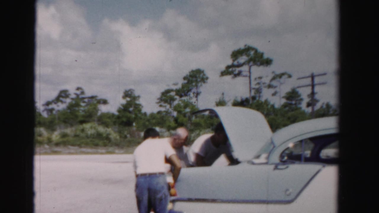 Three men stand beside an open trunk of a light-colored sedan in a sunny roadside lot.