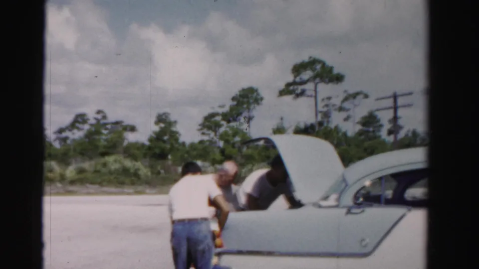 In 1955 Dade County, Florida, three men unload items from the open trunk of a pale 1950s sedan near roadside trees.