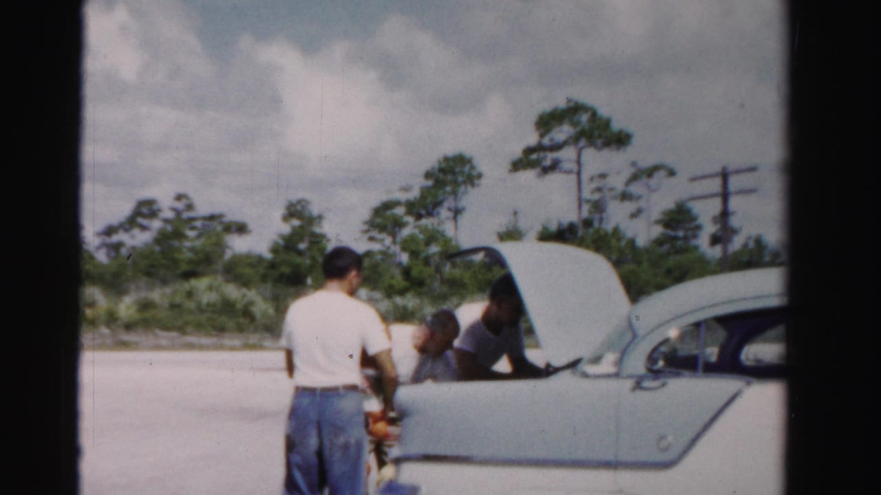 Men gather at the open trunk of a pale sedan, handling groceries near a roadside.