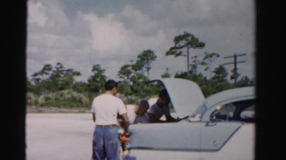 Three men continue unloading groceries from a light-colored 1950s sedan in Dade County, Florida, with trees and utility poles behind them.