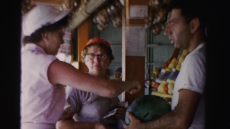At a shaded Dade County produce stall, two women and a man examine or handle produce, including a watermelon.