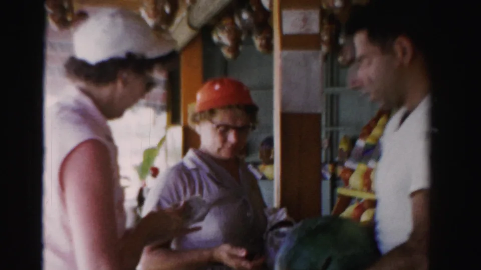 In the 1955 Florida market scene, a woman in a red cap holds produce near another woman and a man by a watermelon.