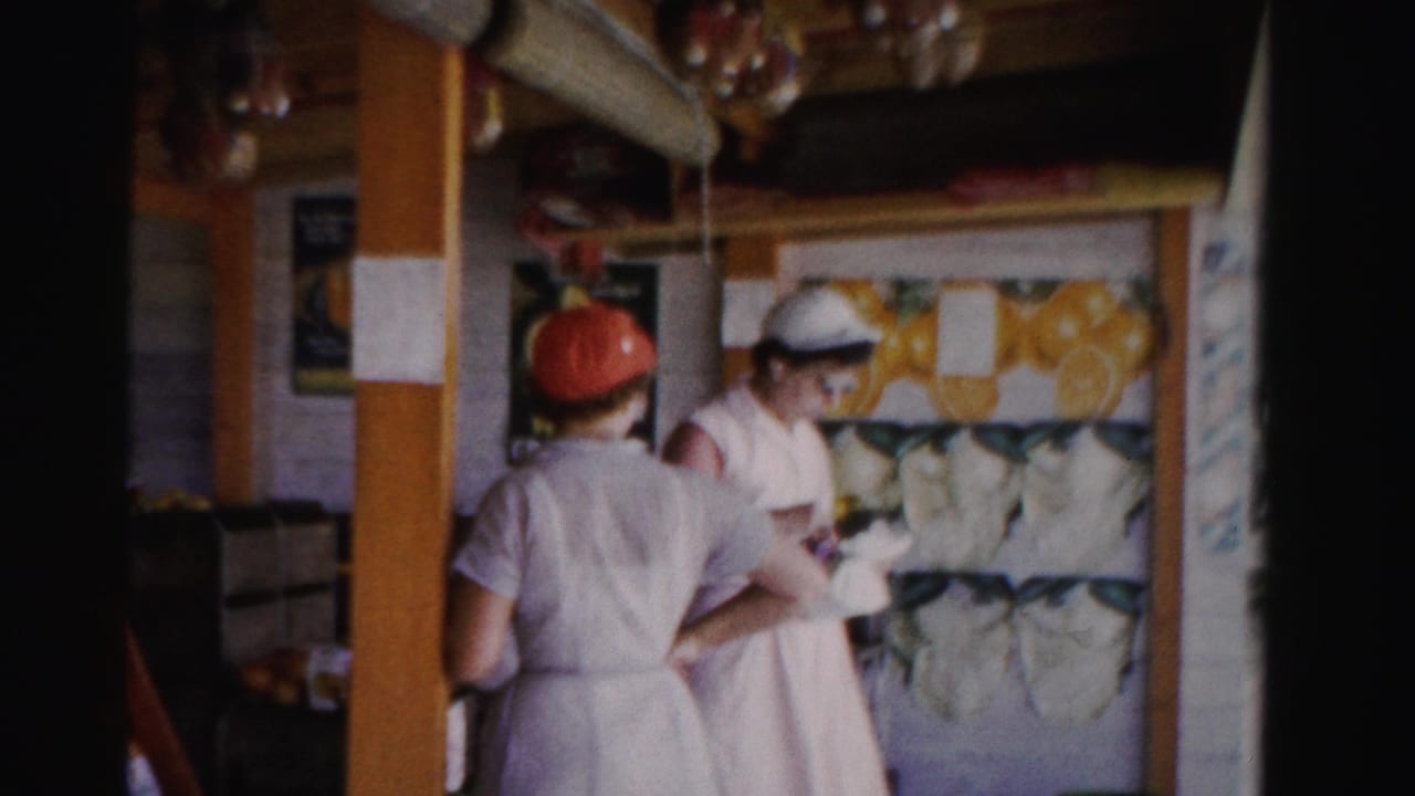1955 Florida fruit-stand frame with two women in white uniforms, one in a red hat and one in a white hat, near produce displays.