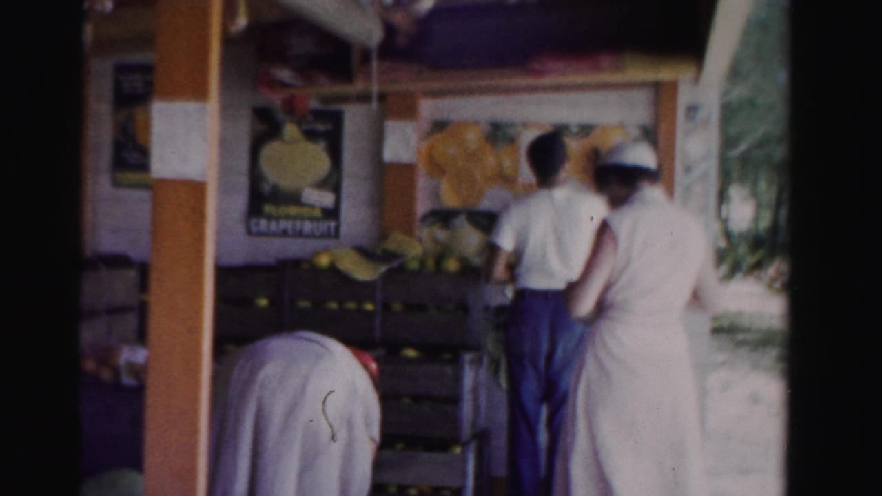 1955 Dade County, Florida frame showing adults near wooden citrus crates at a roadside stand with a Florida grapefruit poster visible.