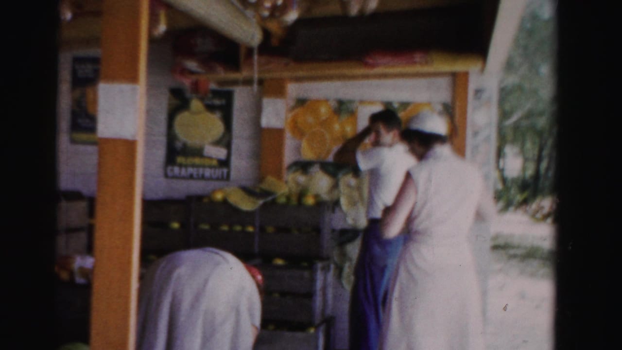 1955 Dade County, Florida frame of adults in light clothing at a citrus stand, with crates and a Florida grapefruit poster in the background.