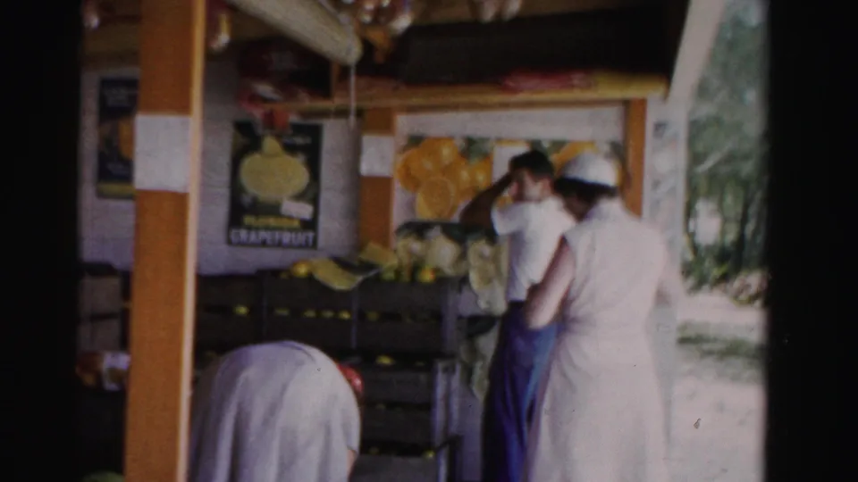 1955 Dade County, Florida frame of adults in light clothing at a citrus stand, with crates and a Florida grapefruit poster in the background.