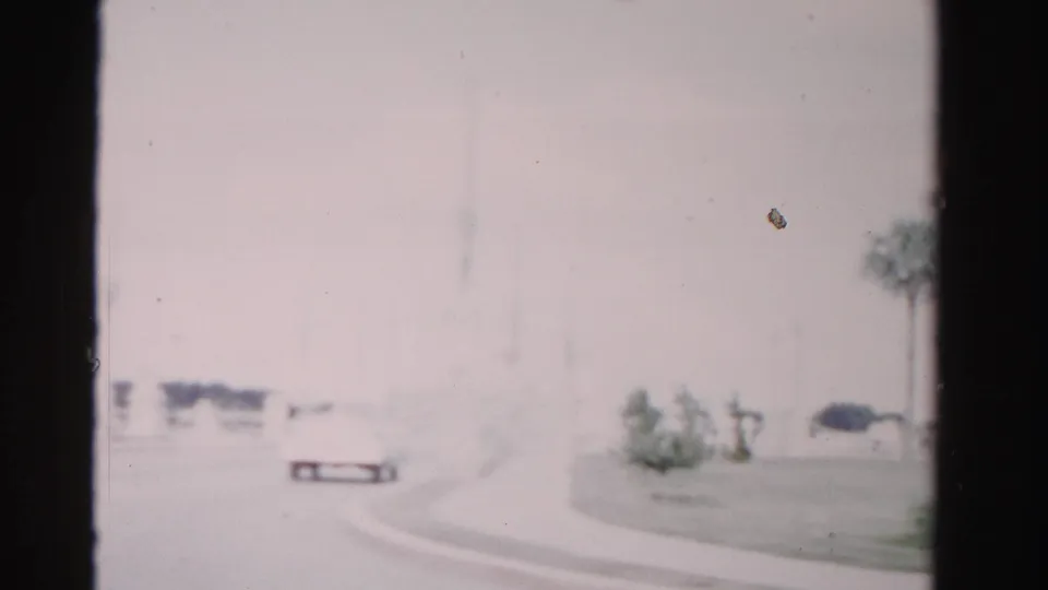 1955 Dade County driving frame with a curving road, a small white car, a palm tree, and a tall vertical marker or pole in a washed-out scene.