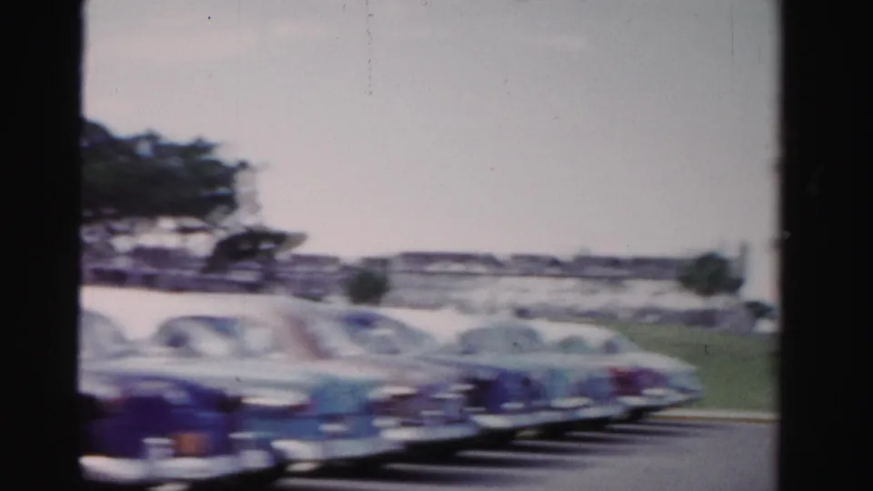 1955 Dade County frame showing several parked rounded American cars beside grass, shade trees, and low roadside buildings.
