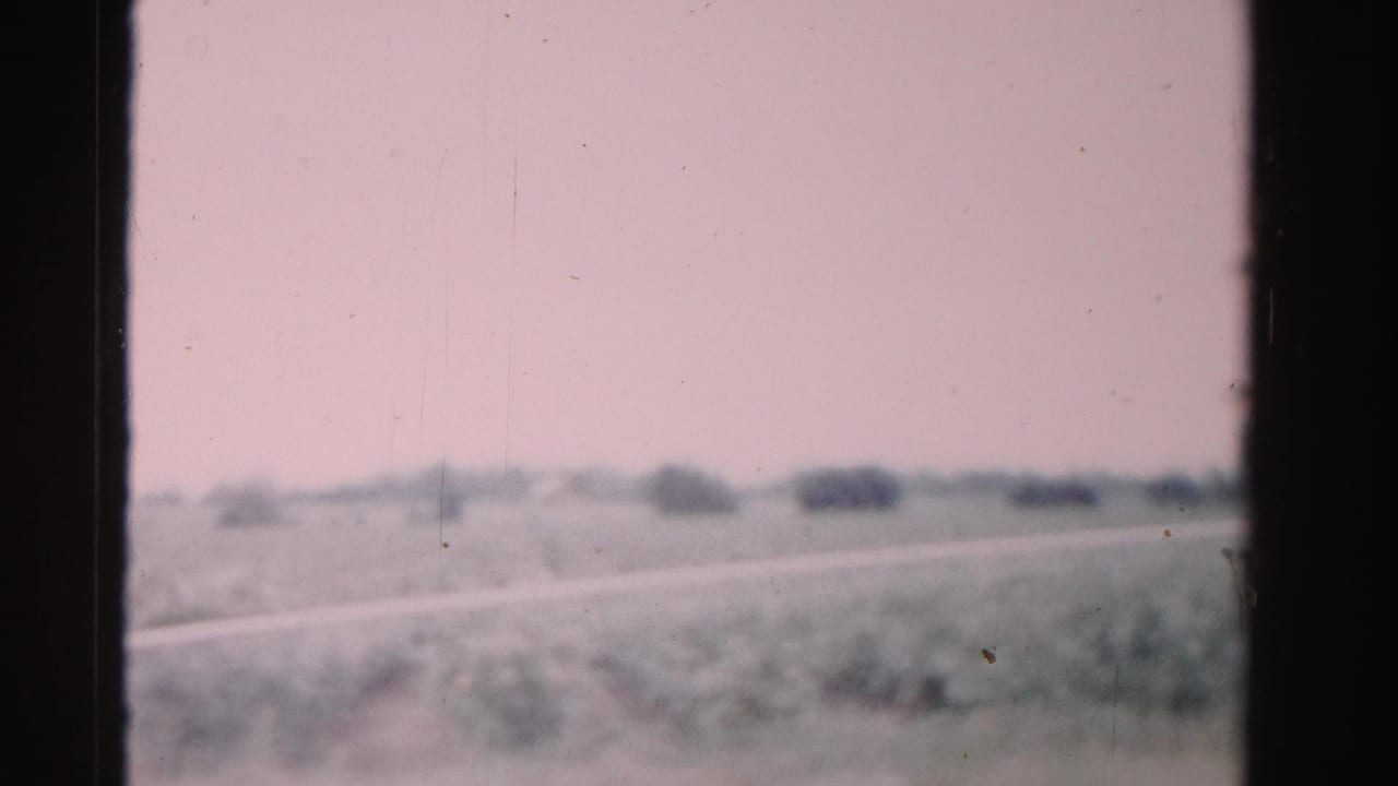 Dade County rail-travel view with soft fields, distant shrubs, a pale horizontal strip, and dark framing from the train window or film gate.
