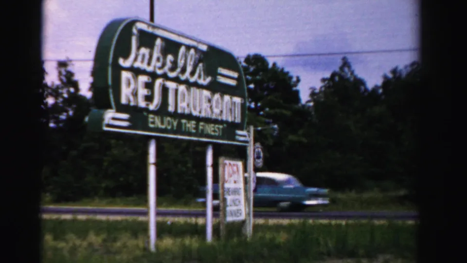 1955 Dade County frame showing Jakella's Restaurant sign, roadside grass, trees, and a passing mid-century car.