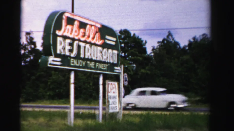 1955 Dade County 8mm frame of the Jakella's Restaurant sign beside a road as a white mid-century car passes.