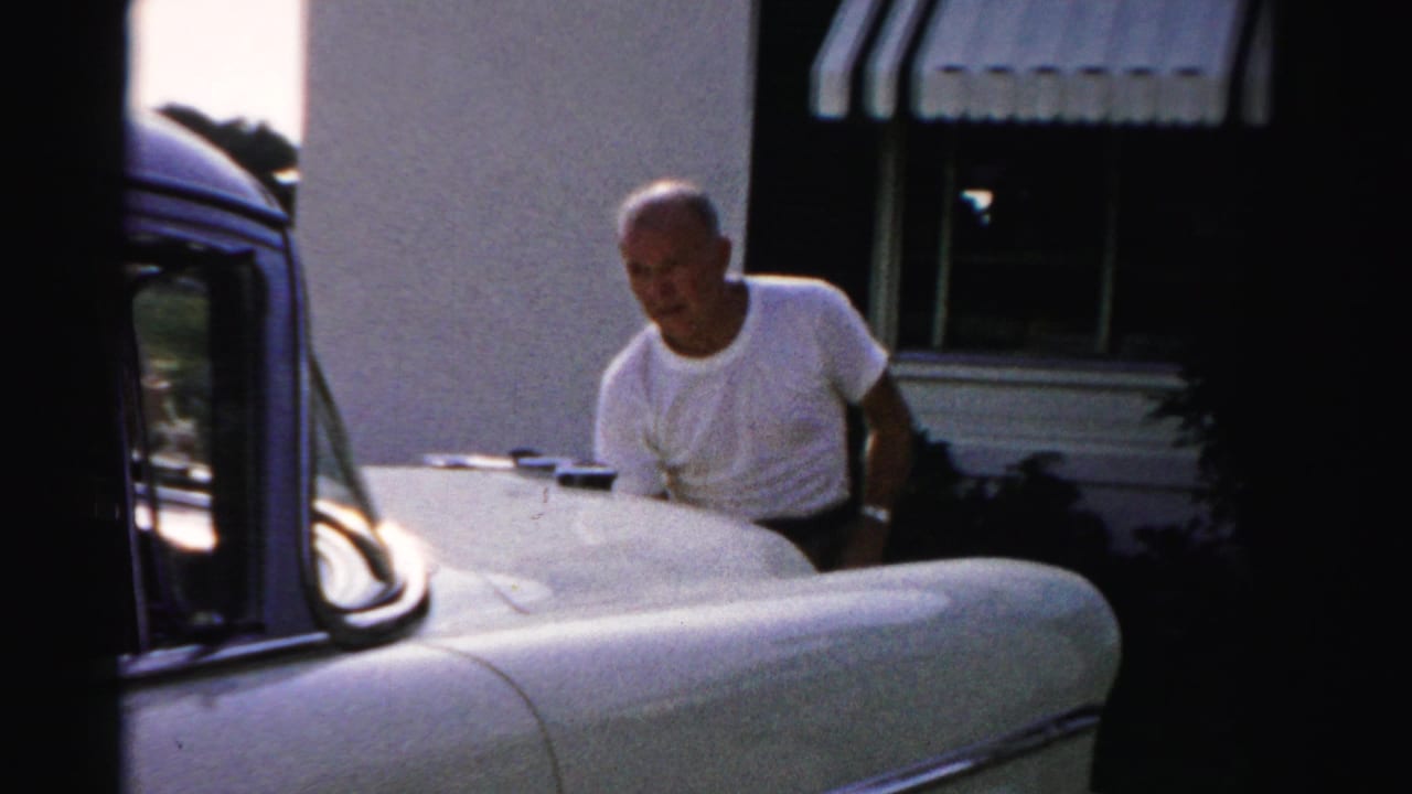 In 1955 Dade County footage, a balding man in a white T-shirt pauses beside a vintage car outside a house with striped awnings.