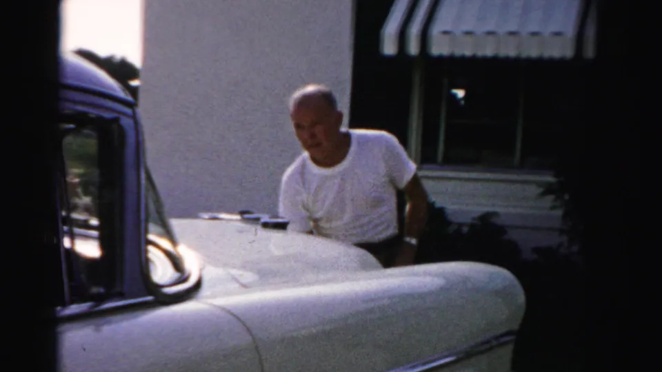In 1955 Dade County footage, a balding man in a white T-shirt pauses beside a vintage car outside a house with striped awnings.