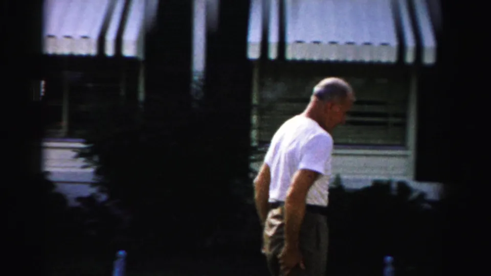 The man walks along the exterior of a Dade County house, with striped awnings, windows, and shrubs visible behind him.
