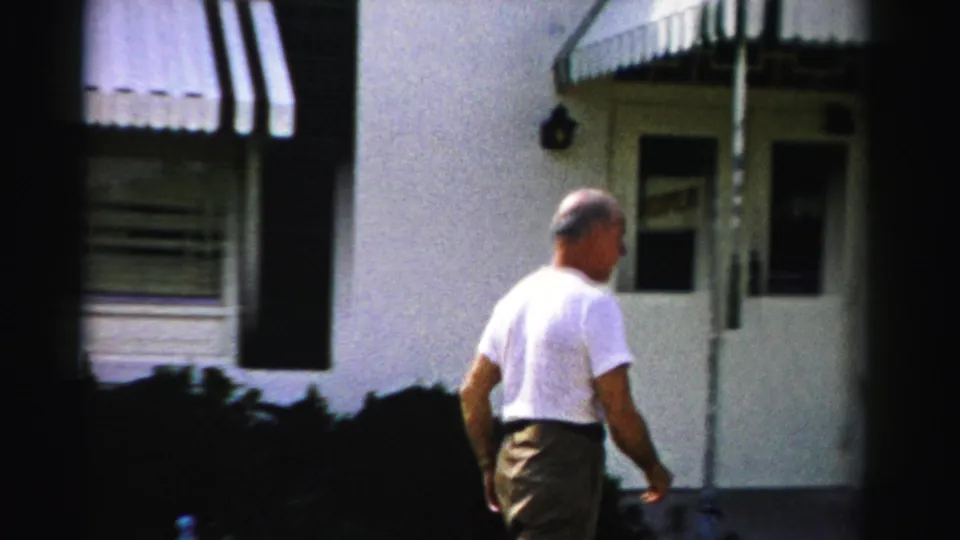 The man continues beside the residential exterior, moving toward a doorway under striped awnings in the 1955 Dade County home-movie scene.