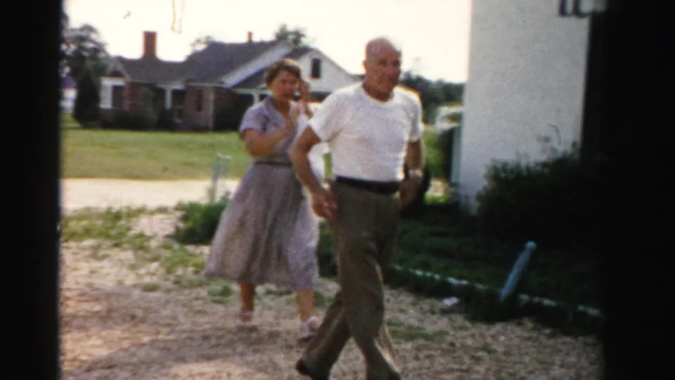 A woman in a light dress follows the man across a yard beside the house, with another residence visible in the background.