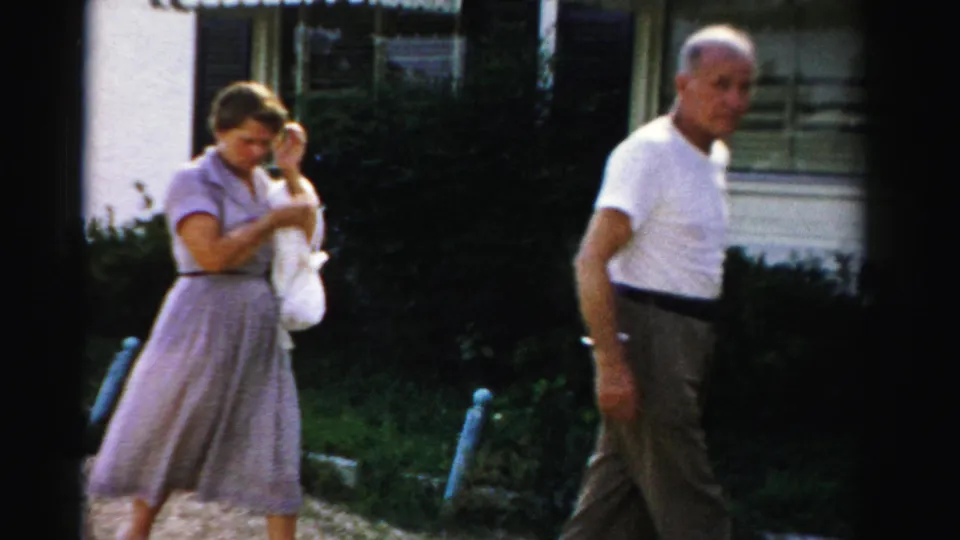 A woman in a lavender dress holds a cloth outside a Dade County suburban home as a man in a white T-shirt walks nearby.