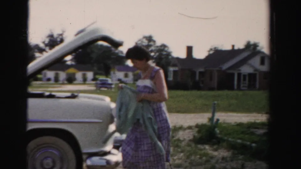 A woman in a patterned dress stands beside a white mid-century car with the hood raised in a 1955 Dade County neighborhood.