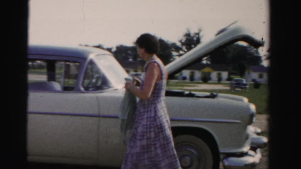 The woman stands beside the white car's raised hood, holding a cloth while suburban houses and lawns sit in the background.