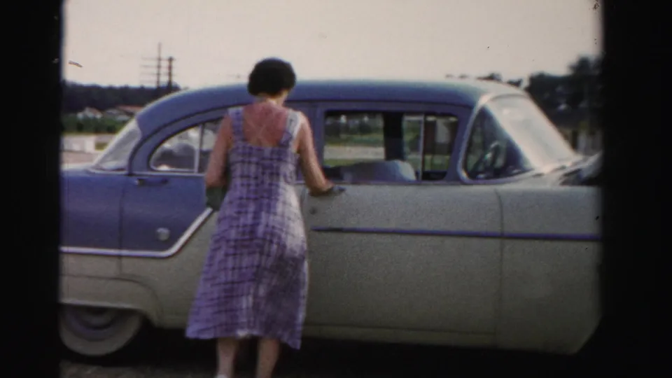 1955 Dade County frame with a woman in a plaid dress facing a two-tone sedan in a roadside setting.