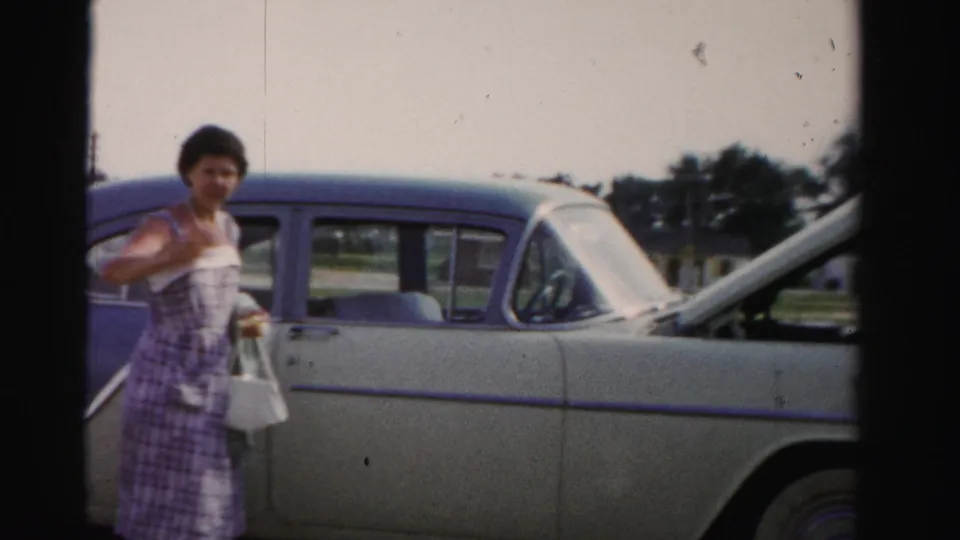 1955 Dade County 8mm still showing a woman turning toward the camera near a two-tone sedan with a bag in hand.