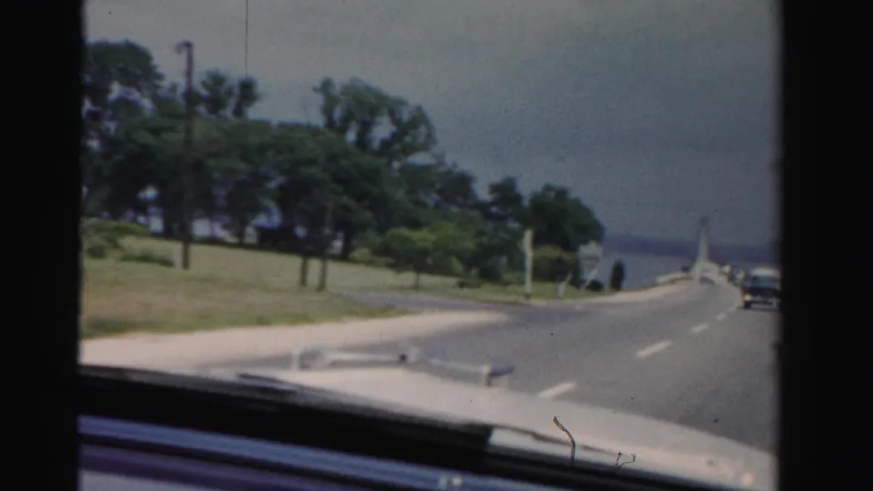 1955 Dade County road view from a moving or parked car, with hood visible, trees to the side, and vehicles in the distance.