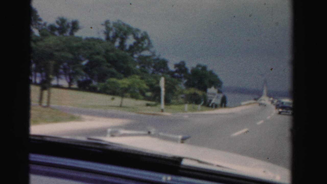 Passenger-seat view of a car approaching a bridge beside trees and water.