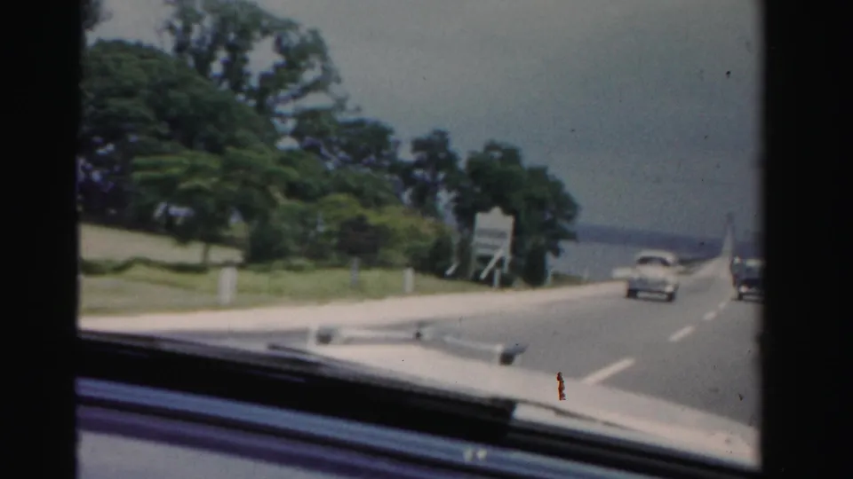 Passenger view in Dade County, Florida shows a mid-century road curving toward a bridge over water, with trees and traffic ahead.