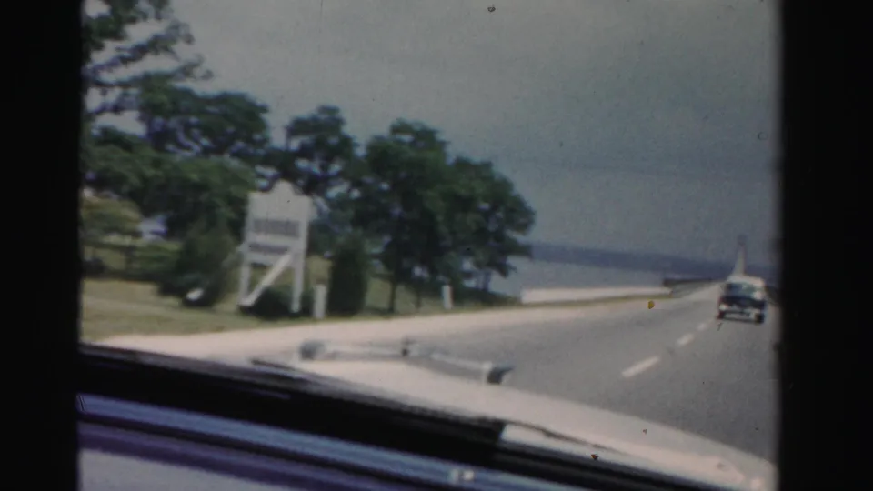 The 1955 home-movie view shows the car nearing a bridge approach, with a roadside sign, trees, water, and a vehicle ahead.