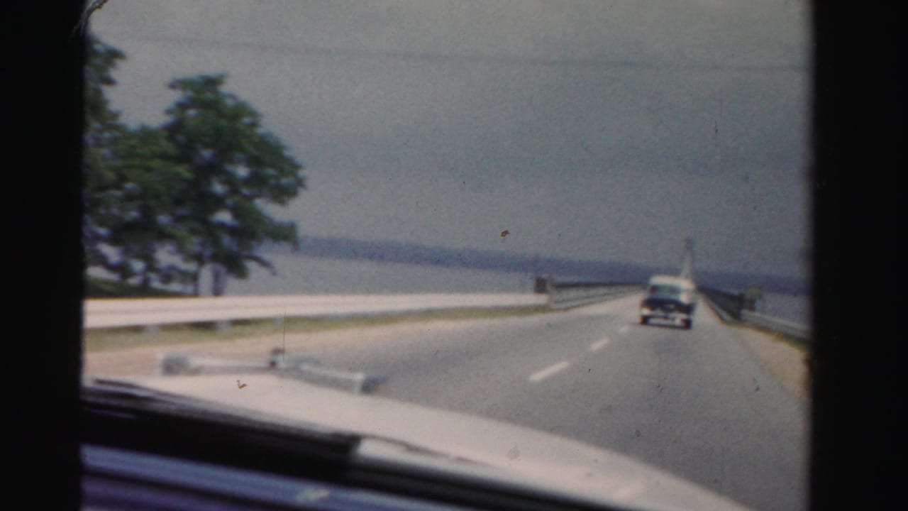 Windshield view of a two-lane bridge road with a car ahead, water at left, trees, and the car hood visible.