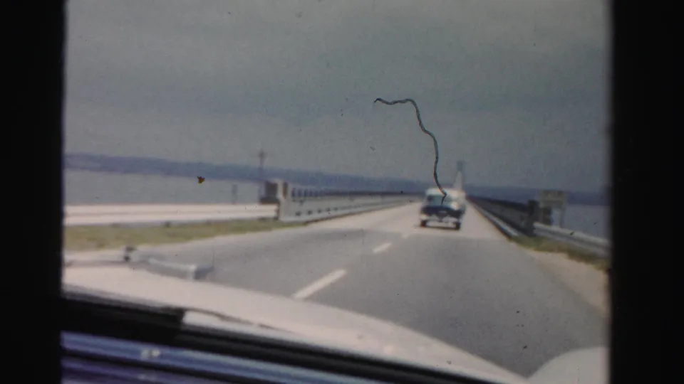 1955 Dade County, Florida road-trip frame looking through a windshield at a long bridge, guardrails, water, and a car traveling ahead.