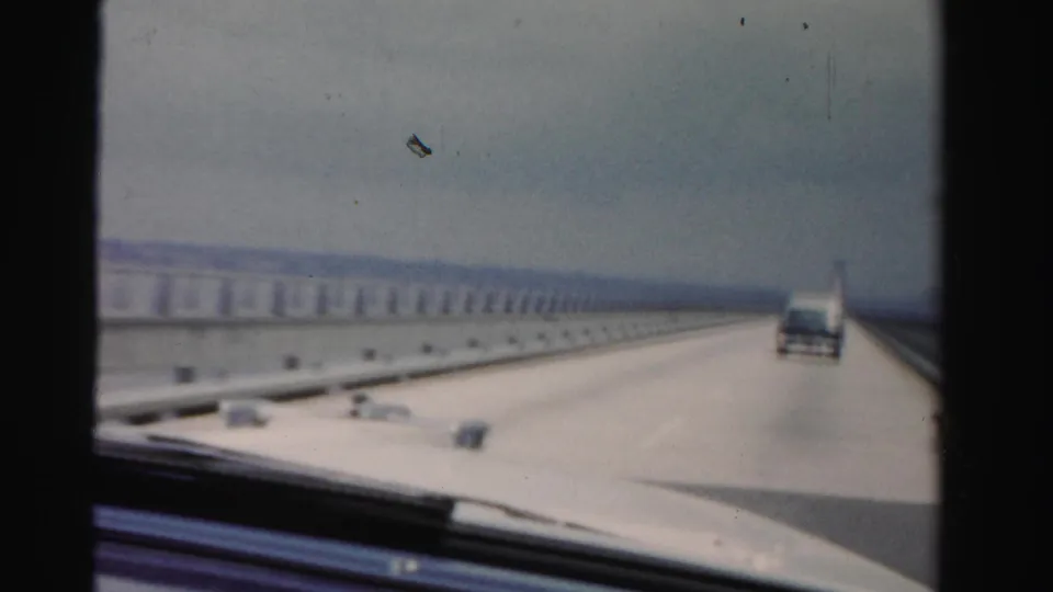 Soft 8mm frame from 1955 Dade County, Florida showing bridge travel from inside a car, with water, railings, and a vehicle ahead.