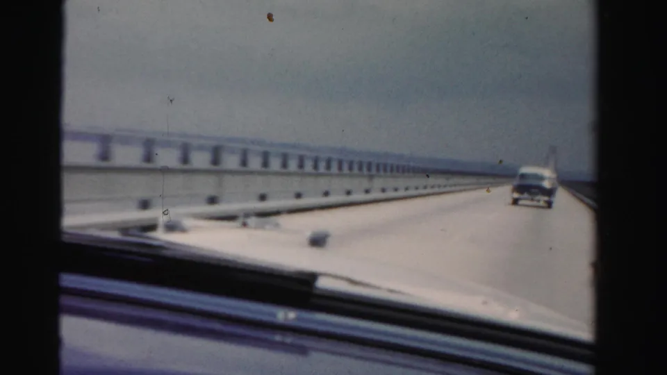 1955 Dade County, Florida 8mm frame showing a forward windshield view on a long bridge, with a car ahead and water beyond the railings.