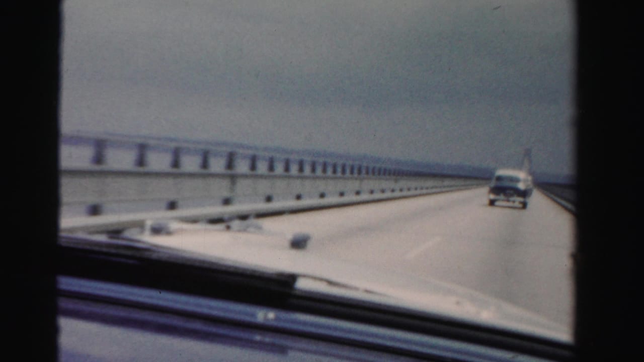 Dashboard view of a car driving onto a long bridge with another vehicle ahead.