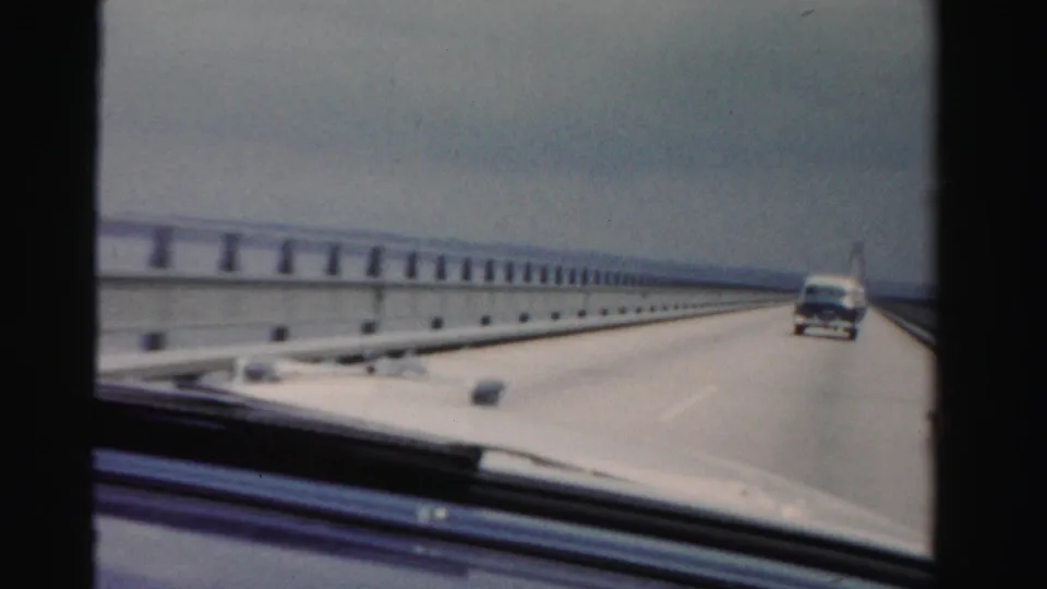 1955 Dade County 8mm dashboard view showing a car traveling along a long bridge with guardrails, water, and a vehicle ahead.