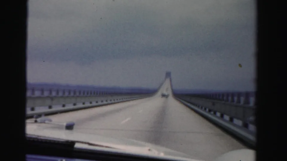 Dashboard perspective in 1955 Dade County shows a long bridge span continuing across water beneath a cloudy sky.